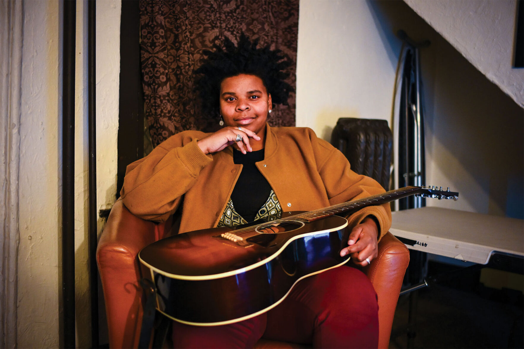 Joy Clark press photo artist seated in leather chair smiles and holds her guitar